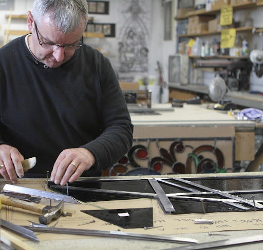This is Patrick who is piecing together a stained glass panel. The metal strips are called lead came, which has an H-shape. The glass sections fit snugly into the grooves of the came. Came comes in many widths.